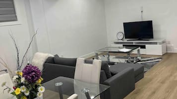 A modern living room with a glass dining table, a sofa, a TV, and a decorative vase with flowers.