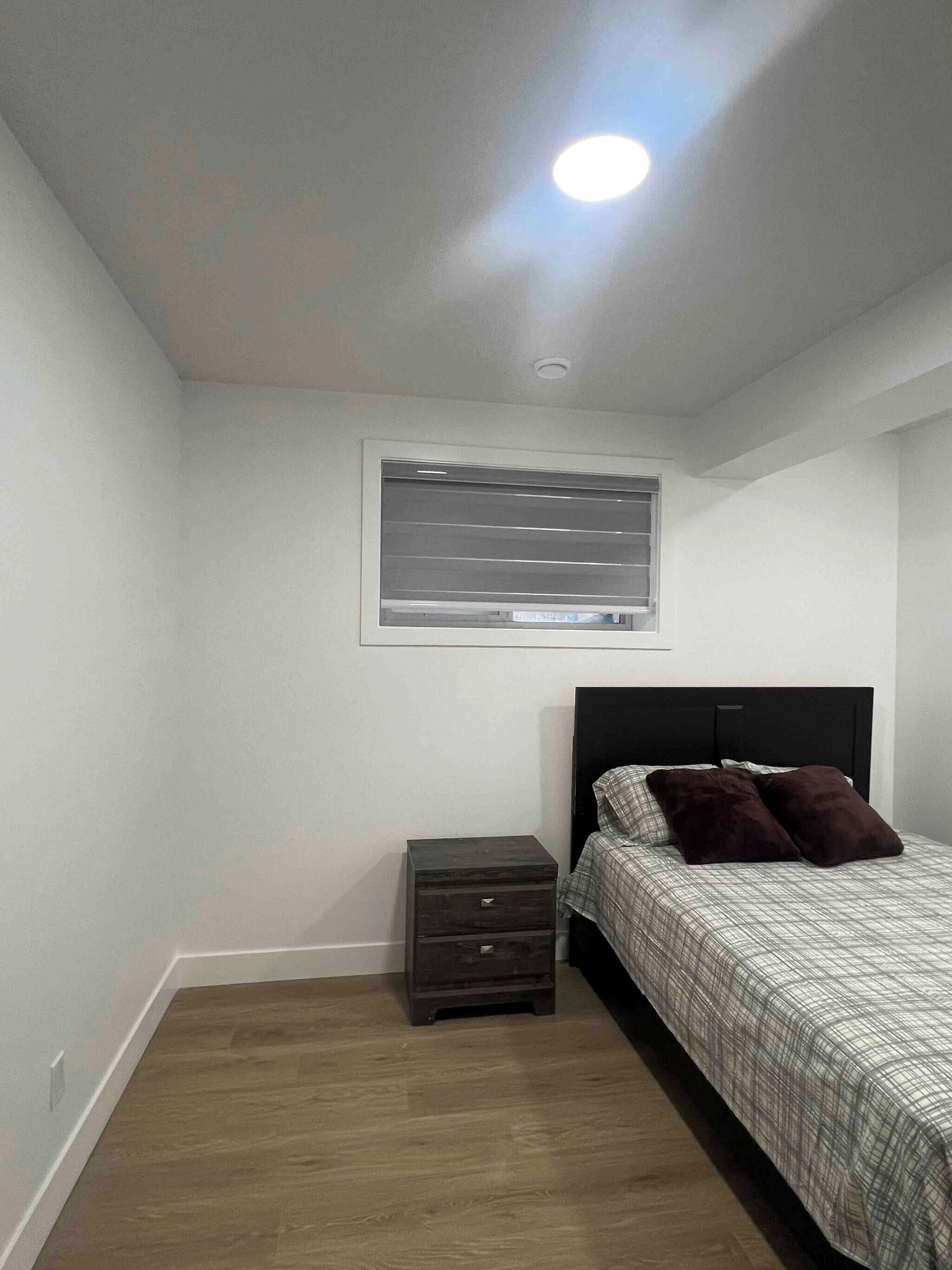 A bedroom with a bed, a nightstand, a small drawer, a window with blinds, and a ceiling light.