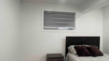 A bedroom with a bed, a nightstand, a small drawer, a window with blinds, and a ceiling light.