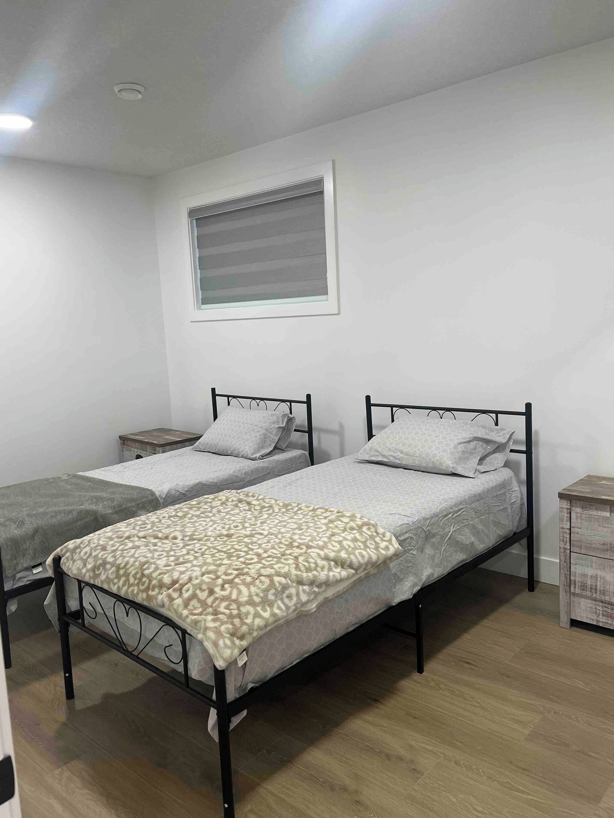 A room with two single beds, a window with blinds, a small bedside table, and a wooden floor.
