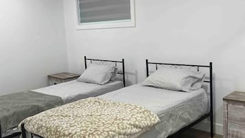 A room with two single beds, a window with blinds, a small bedside table, and a wooden floor.