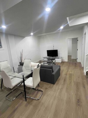 A modern living room with a glass dining table, white chairs, a grey sofa, and a flat-screen TV. - Room-magnificent Brand New Luxurious Bas (Edmonton)