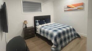 A bedroom with a bed, a nightstand, a lamp, a TV, and a painting on the wall. - Room-magnificent Brand New Luxurious Bas (Edmonton)