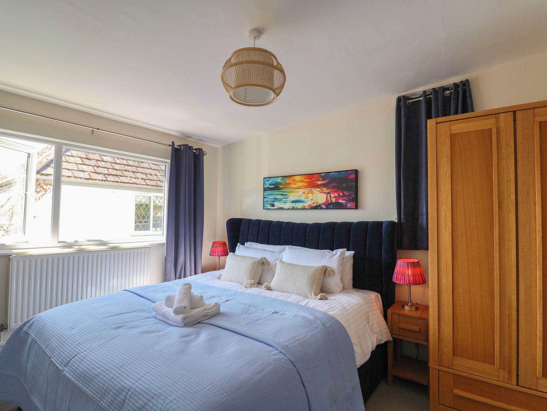 A bedroom with a bed, a window, a painting, a wardrobe, and a bedside lamp.
