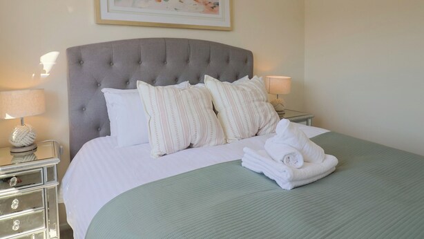 A neatly made bed with a grey upholstered headboard, white pillows, and a green bedspread. There are two bedside tables with lamps and a framed artwork above the bed. - 2 Mill Green Court (Lyme Regis)