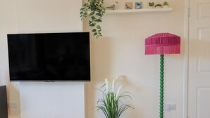 A living room with a flat-screen TV mounted on the wall, a potted plant, a floor lamp, and a small pig figurine on the floor. - 2 Mill Green Court (Lyme Regis)