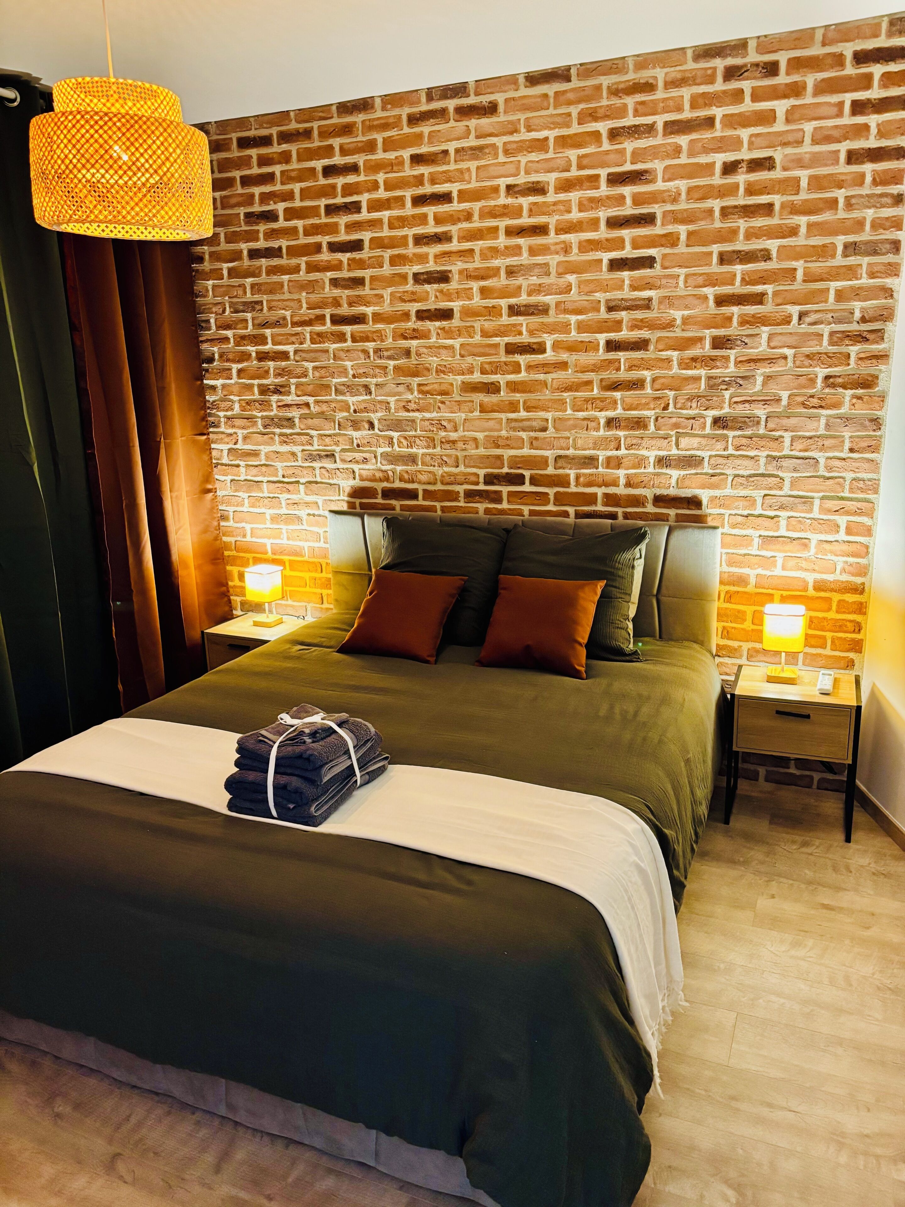 A hotel room with a brick wall, a bed with a green bedspread, two bedside tables with lamps, and a hanging light fixture.