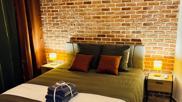 A hotel room with a brick wall, a bed with a green bedspread, two bedside tables with lamps, and a hanging light fixture. - House with a renovated courtyard—"Le Cocon d'Albi"—5 minutes from downtown (Albi)