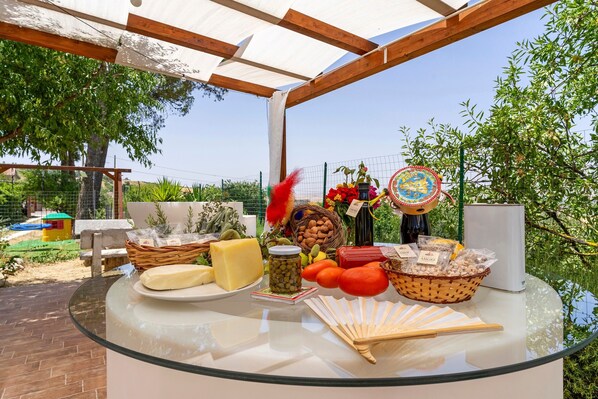 An outdoor dining area with a glass table displaying various food items, a wicker basket, and a decorative fan.