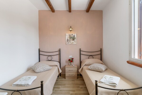 A small, well-lit bedroom with two single beds, a nightstand, a wall-mounted lamp, and a window.