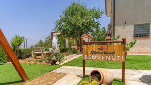 A wooden sign with 'NewLife Villalba Resort BENVENUTI' text, a garden with succulents, and a clear sky.