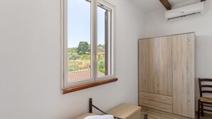 A bedroom with a bed, a wooden wardrobe, a window with a view of greenery, and an air conditioning unit.