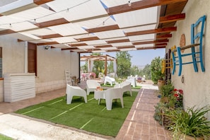 Un patio al aire libre con sillas de plástico blanco, una mesa pequeña con un vaso y una pérgola con luces colgantes.