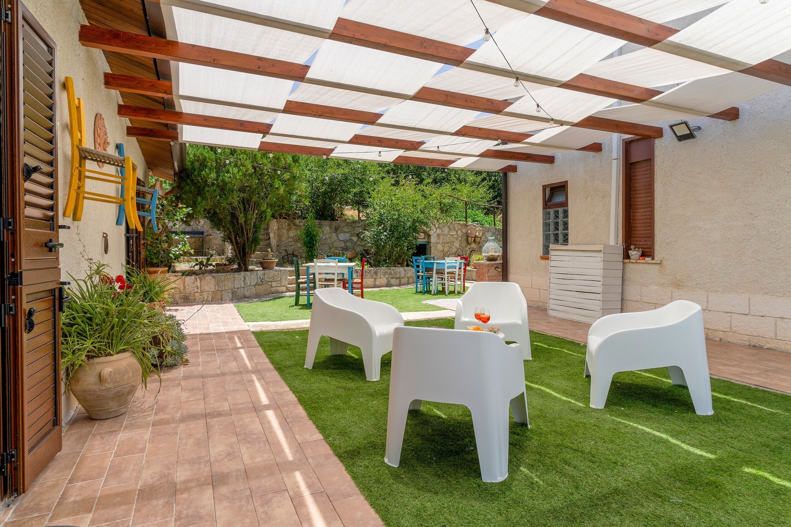 Une terrasse avec des chaises et une table en plastique blanc, une pergola surmontée d’une toile blanche et, au fond, un mur de pierres.