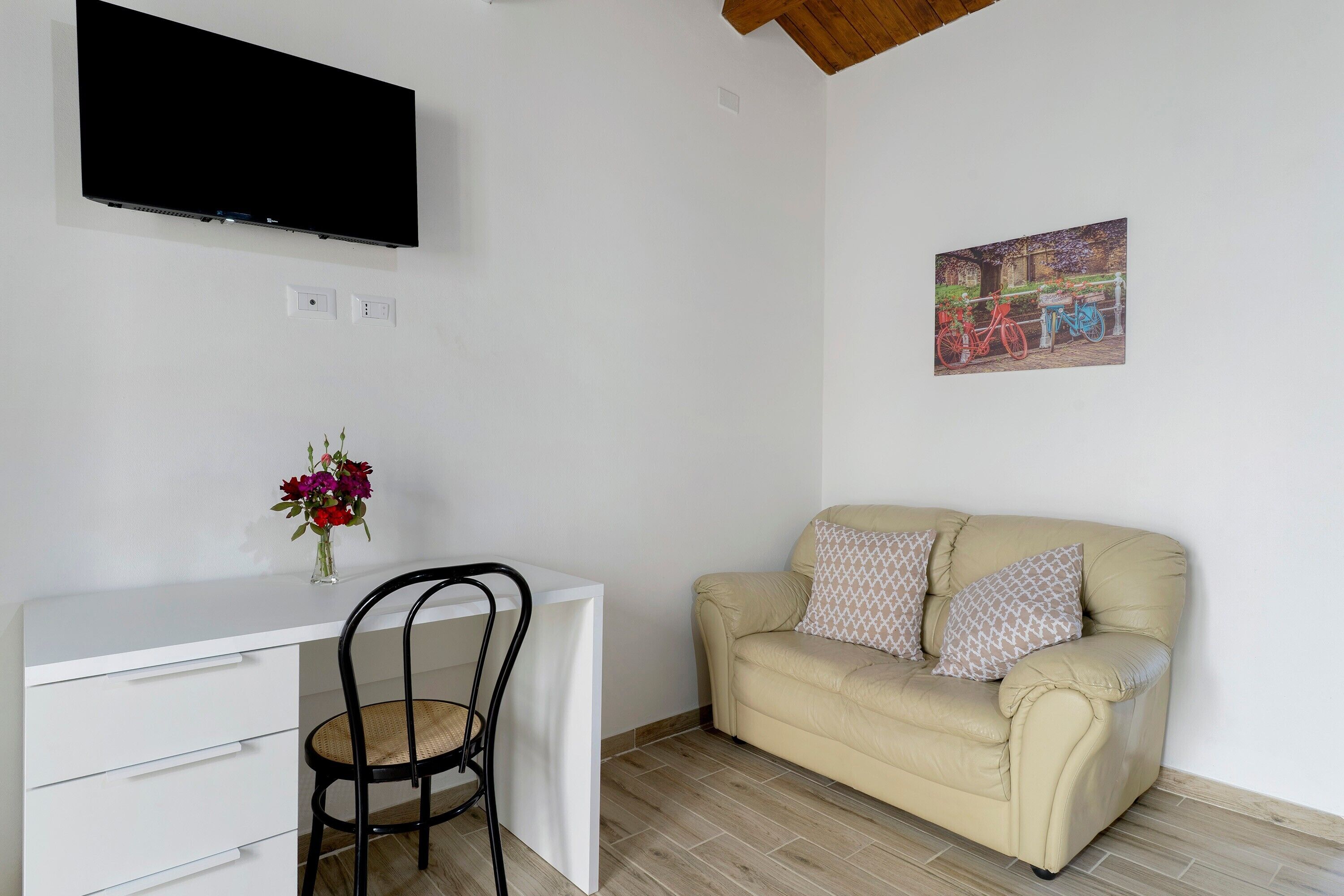 Une pièce avec un bureau blanc, un canapé beige agrémenté de coussins à motifs, un téléviseur fixé au mur et un tableau encadré accroché au mur.