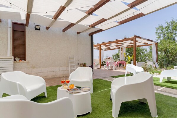 An outdoor patio area with white plastic chairs, a small table with drinks, and a pergola with a white cloth.
