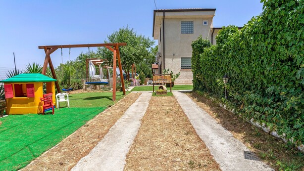 A children's play area with swings and a small hut, a paved pathway, and a building with a sign in the background. - NEW LIFE Villalba Resort - Room '101' with Shared Terrace, Wi-Fi and Air Conditioning (Villalba)