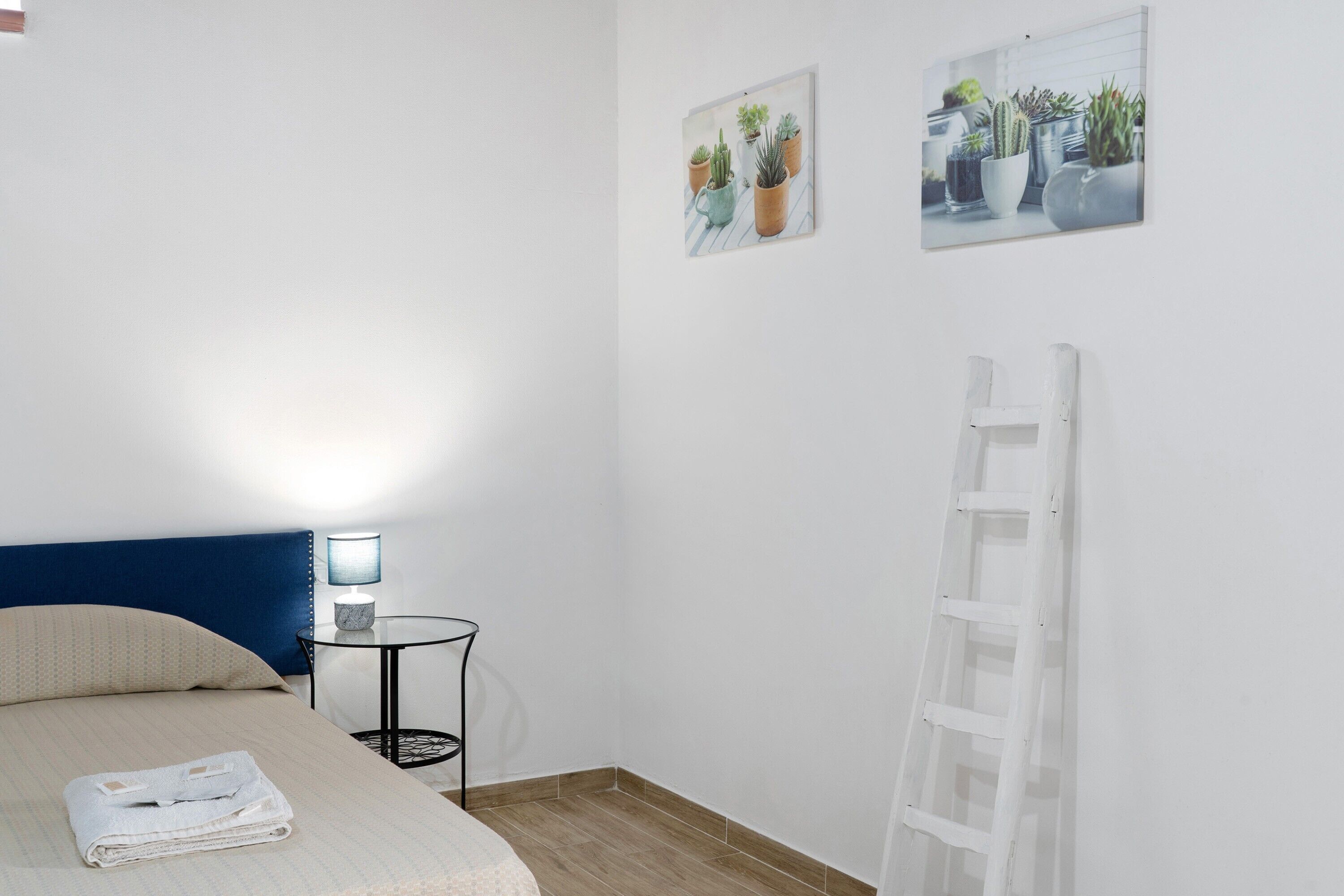 Ein Schlafzimmer mit einem Bett, einem Nachttisch mit Lampe, einer weißen Hakenleiter und zwei Pflanzenbildern an der Wand.