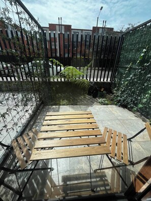 Una panchina in legno con ringhiere in metallo, un piccolo giardino con piante e vista su edifici sullo sfondo.