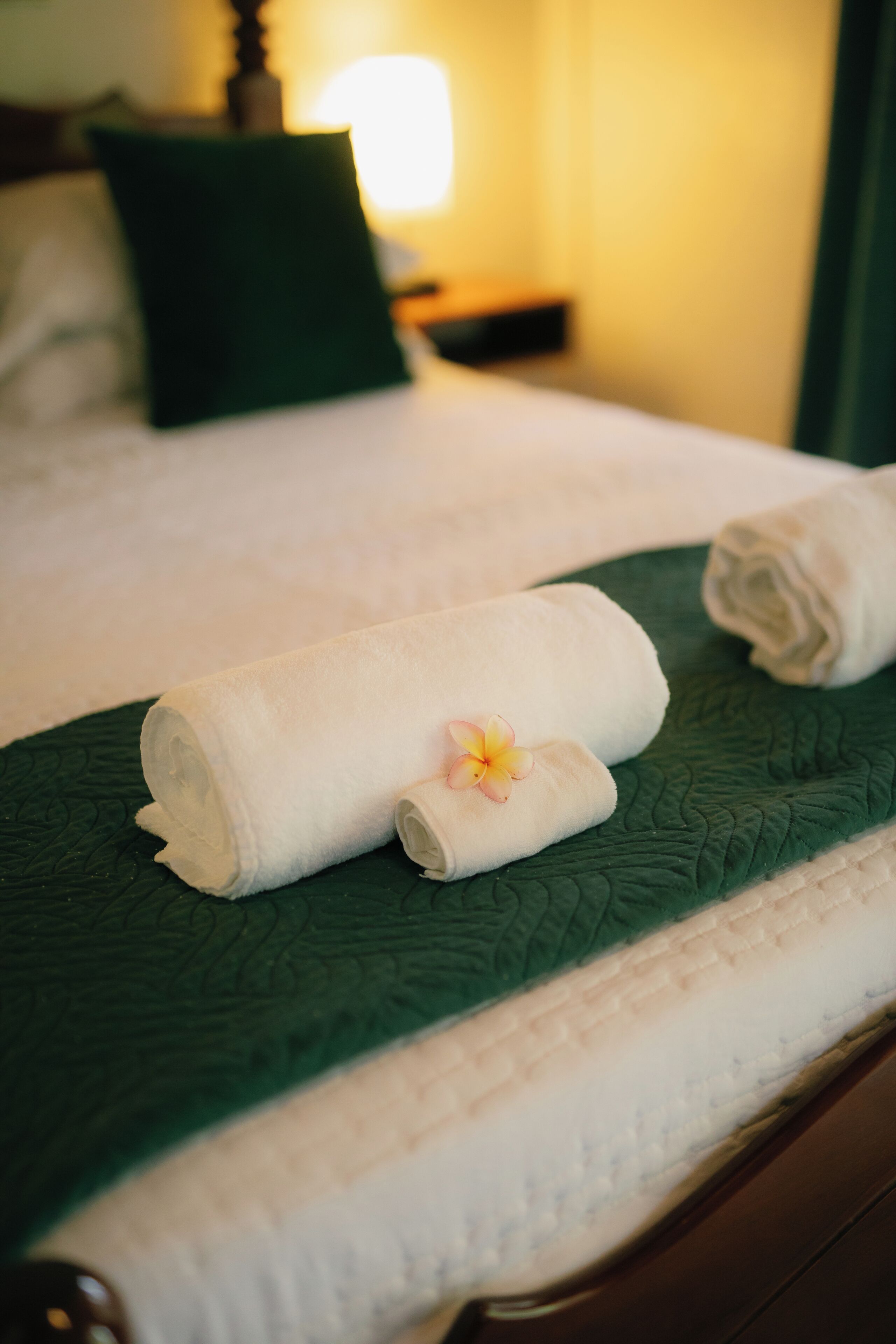 A neatly made bed with a green bedspread, a rolled white towel, and a single flower.