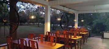 Un espacio para comer al aire libre con mesas y sillas de madera, ruedas grandes como decoración, y luces colgantes.
