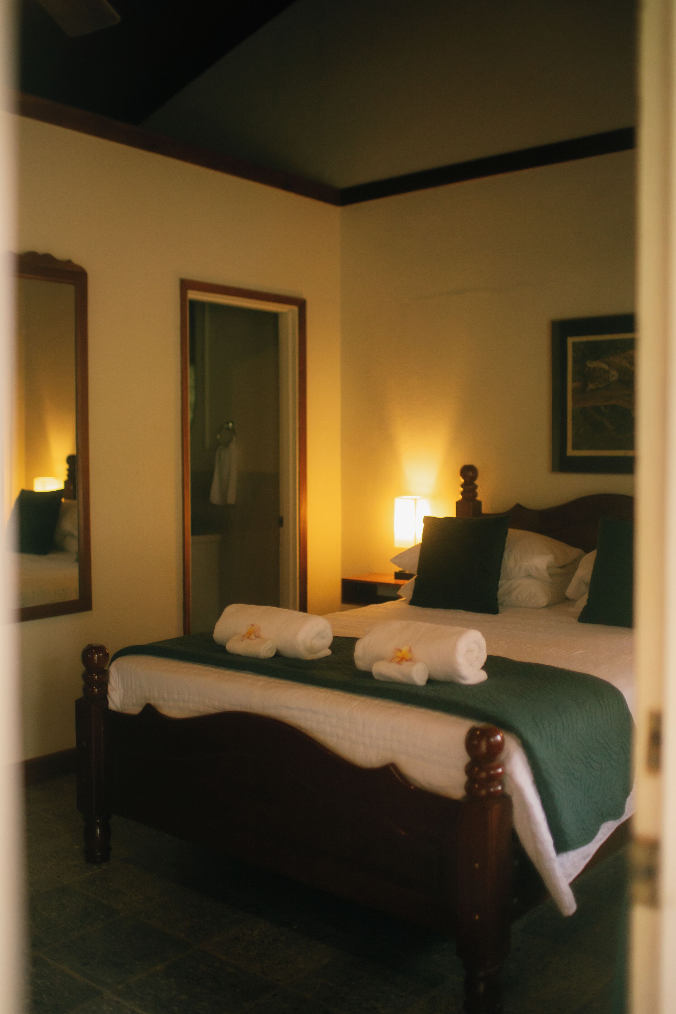 Una cama bien hecha con una manta verde y dos toallas enrolladas, una lámpara de noche, un cuadro en la pared y un espejo.