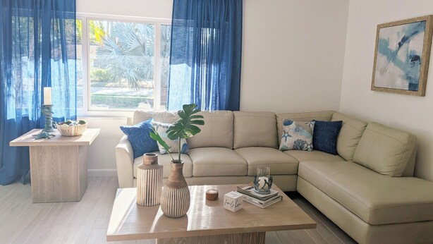 A living room with a beige sofa, a wooden coffee table, and a window with blue curtains. - Siesta Breeze (Sarasota)