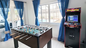 A foosball table and a Midway arcade machine in a room with large windows and blue curtains. - Siesta Breeze (Sarasota)