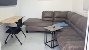 A modern living room with a brown leather sectional sofa, a wooden desk, a black office chair, and a small table with a remote control. - Siesta Breeze (Sarasota)