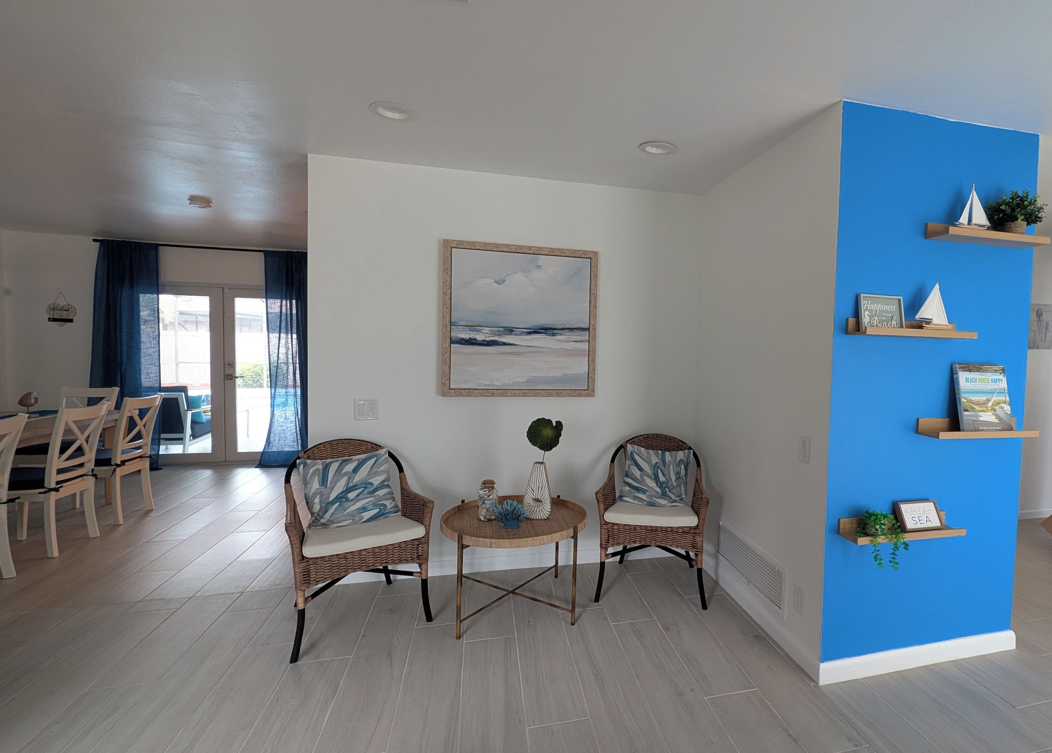 A living room with two wicker chairs, a round coffee table, a painting, and a blue accent wall with shelves.