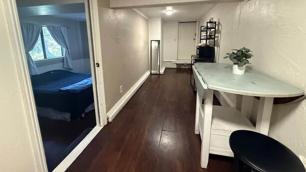A narrow hallway with a bed on the left, a white table with a chair on the right, and a door at the end.