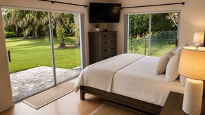 Un dormitorio con cama, televisor, cómoda y vista a un jardín a través de amplios ventanales correderas.