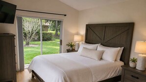 Un dormitorio con una cama grande, un ventanal corredero a un jardín, un televisor montado en la pared y una mesita de noche con una lámpara.