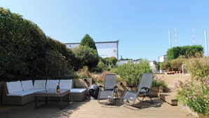 Ein Sitzbereich im Freien mit einer Couch und Liegestühlen, umgeben von viel Grün und blauem Himmel.