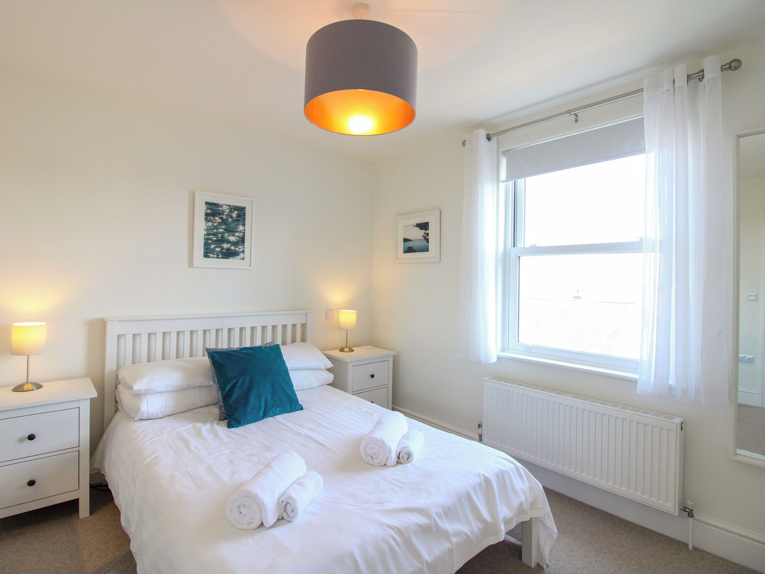 Een netjes opgemaakt bed met witte lakens en opgerolde handdoeken, twee nachtlampjes, een witte nachtkastje, een raam met witte gordijnen, en een wandlamp.