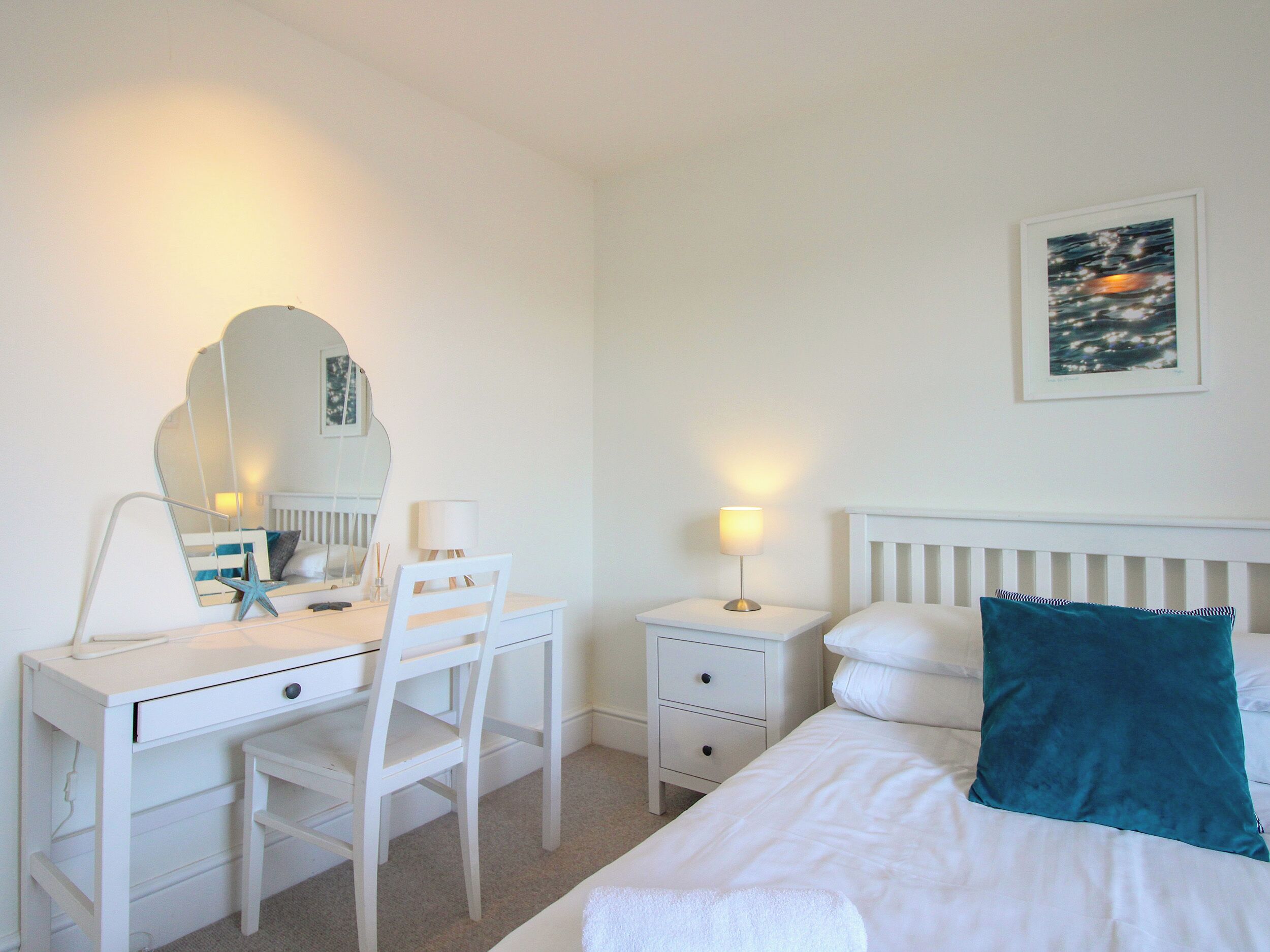Habitación de hotel con cama, escritorio y silla, mesita de noche con lámpara, mesita auxiliar, cómoda y espejo adosado a la pared.