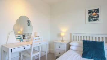 Habitación de hotel con cama, escritorio y silla, mesita de noche con lámpara, mesita auxiliar, cómoda y espejo adosado a la pared.