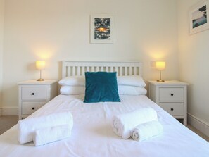 A neatly made bed with white linens, a teal pillow, and rolled towels on a white headboard. - Lorrell (Salcombe)