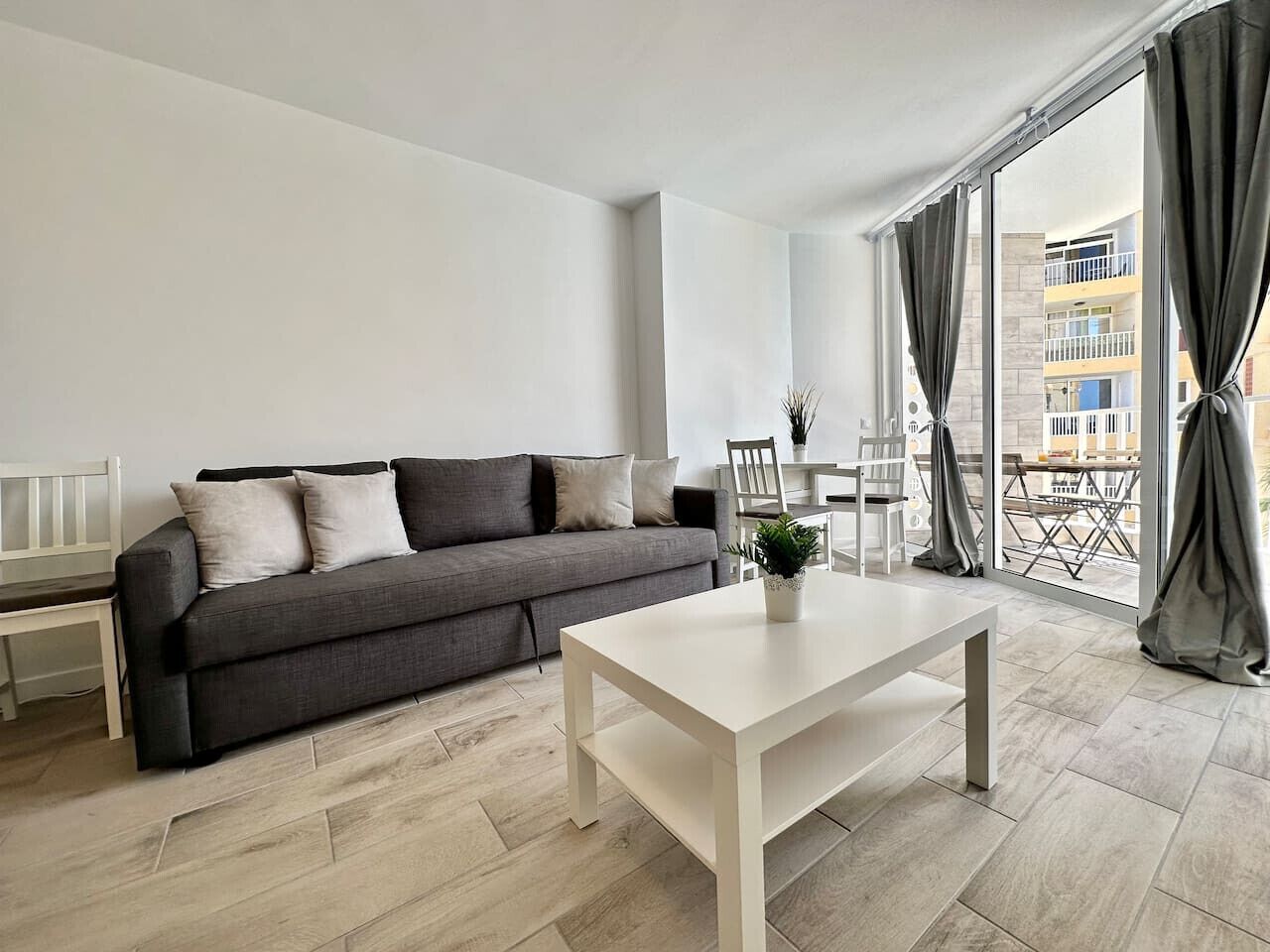 Una sala moderna con un sofá gris, una mesa de centro blanca y un balcón con cortinas.