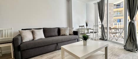 Una sala moderna con un sofá gris, una mesa de centro blanca y un balcón con cortinas.