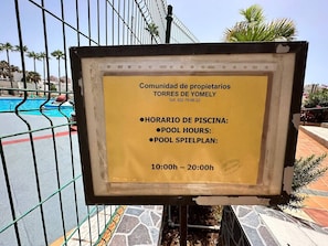 A sign with pool hours and a pool area in the background. - Yomely Towers - Y4G (Playa de las Américas)