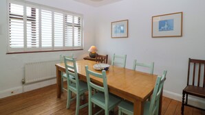 Een eetkamer met een houten tafel, zes stoelen, een raam met jaloezieën, een radiator en twee ingelijste foto's aan de muur.