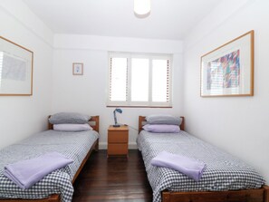 Ein Zimmer mit zwei Einzelbetten, einem Nachttisch, einem Schreibtisch, einem Fenster mit Jalousien und gerahmten Bildern an den Wänden.