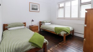 Een kamer met twee bedden, een nachtkastje, een lamp, een lijst met een foto aan de muur en een raam met jaloezieën.