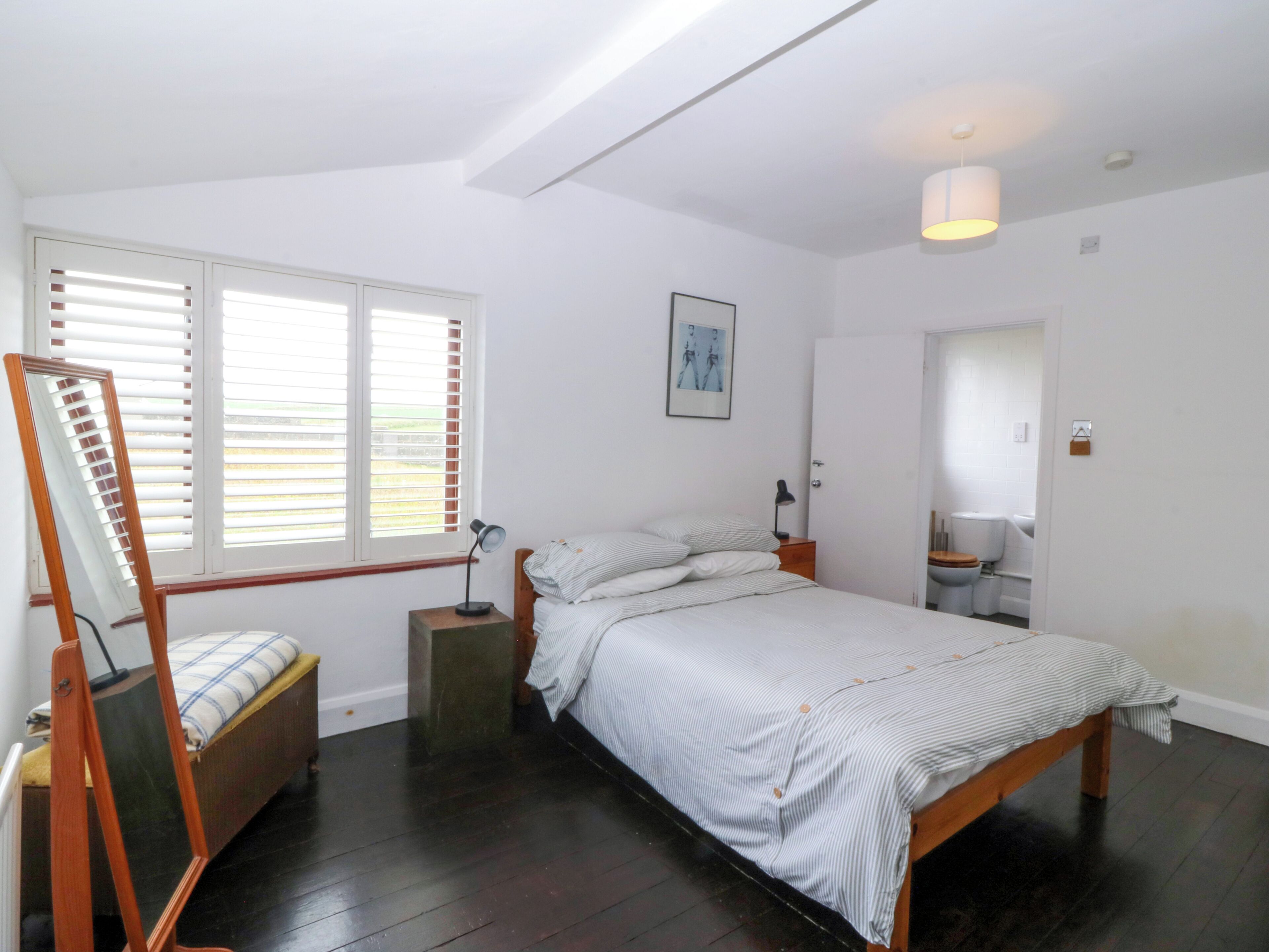 Un dormitorio con cama, una mesita de noche, lámpara, espejo, ventana con persianas y puerta que da a un baño.