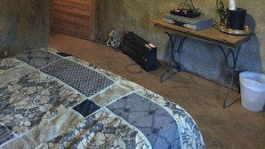 Una habitación rústica con una cama, un televisor, una mesita con dispositivos electrónicos y una puerta de madera.