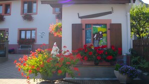 A building with a white facade, a wooden door, and a window with shutters. There are flower boxes with red flowers and a bench outside. - Charming Cottage at the Foot of the Rock (Sewen)