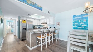 A modern kitchen with white cabinetry, a central island with bar stools, and a dining area with a table and chairs. - Perfect family stay by Gulf & Lagoon! Dogs welcome (Gulf Shores)