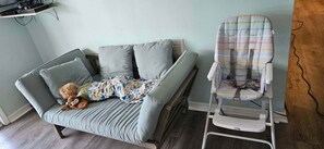Une pièce avec un canapé gris, un transat pour bébé et un ours en peluche.