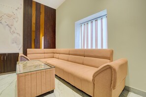 A room with a beige sofa, a glass coffee table, a wooden panel wall, and a window with vertical blinds. - Classic Ummed Palace (Mathura)